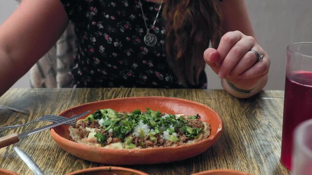 Woman pouring avocado green salsa over traditional Mexican huarache topped with meat, onion and cilantro at food market in Tapalpa, Jalisco, Mexico. Close up shot.