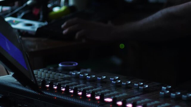 Professional mixing console with illuminated buttons and faders in a dark recording studio. In the background, a sound engineer works at a computer during a music track mixing session