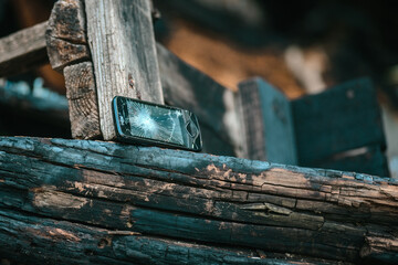 Fototapeta premium Damaged smartphone with spiderweb cracked display lying beside weathered blackened timber from burned barn remains, creating somber close view with rustic textures, smoke stains, and fire aftermath