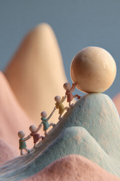 Four small figures work together to push a large round object uphill on a pastel-colored landscape. The scene shows teamwork and effort in a playful setting
