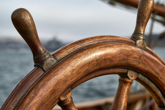A wooden ship's steering wheel is in focus with the sea and distant land behind it. The light shows the warm tones of sunset reflecting off the water
