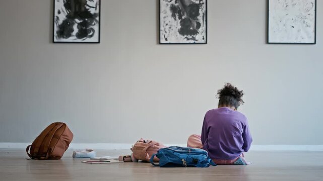 African American child sitting on floor of art gallery with backpacks, seen from rear view. Clip is perfect for educational marketing, museum advertisements, or school field trip campaigns