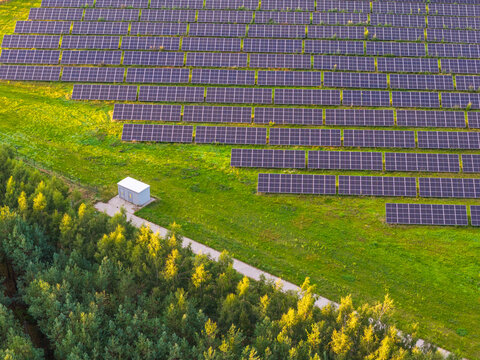 Aerial view of rows of photovoltaic panels in a solar farm surrounded by lush green grass and a forest edge with a small utility building in Gniew, Pomeranian Voivodeship, Poland.