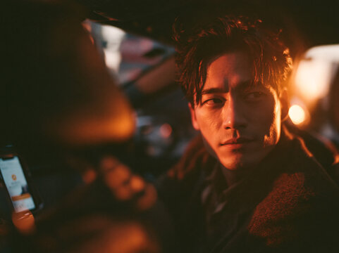 An intense Asian man with messy dark hair and light stubble sits in a car at night, turning to look back over his shoulder toward the passenger seat, his face half-lit by a warm golden glow from outsi