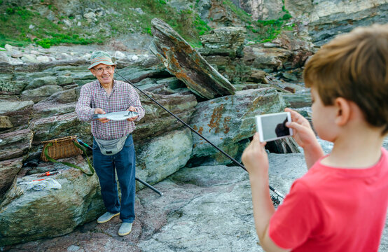 Boy taking picture of smiling grandfather holding fish on fishing line