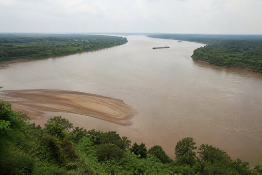 View from high bluff of wide brown river bending through low green forest, sandbar, tugboat and barge downstream, Mississippi River landscape