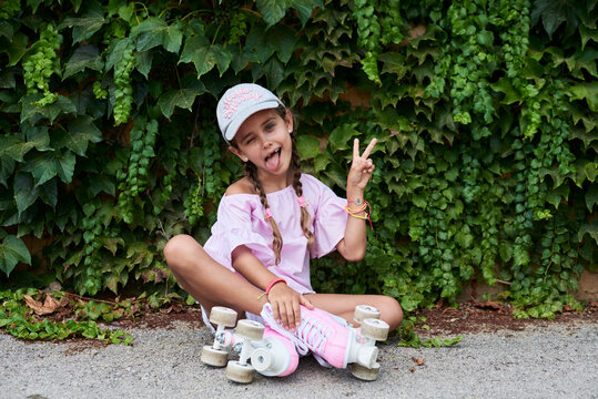 Confident little girl sticking out tongue sitting on the ground with pink roller skates