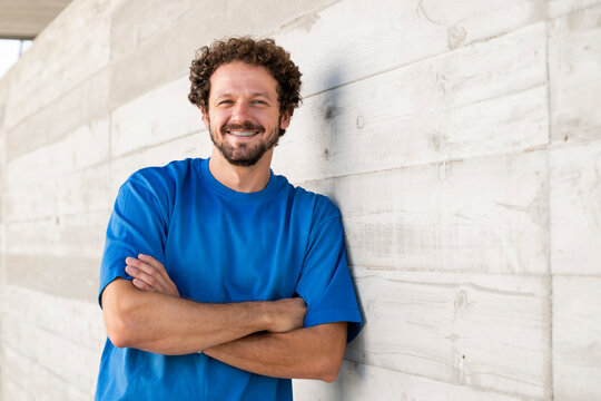Confident man with curly hair leans against a concrete wall outdoors