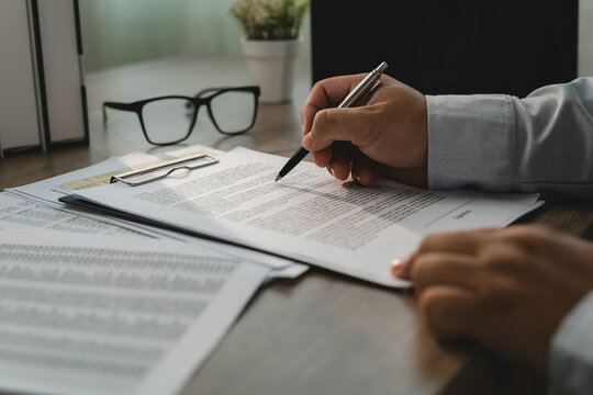Hands holding a pen over printed agreement papers on a clipboard, showing a business person reviewing terms and preparing to sign a contract in an organized office documentation workflow.