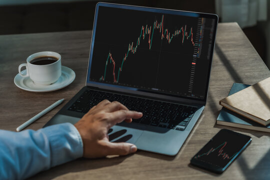 Businessman reviewing candlestick charts on laptop and smartphone on desk, showing online trading analysis, investment research and financial market monitoring.