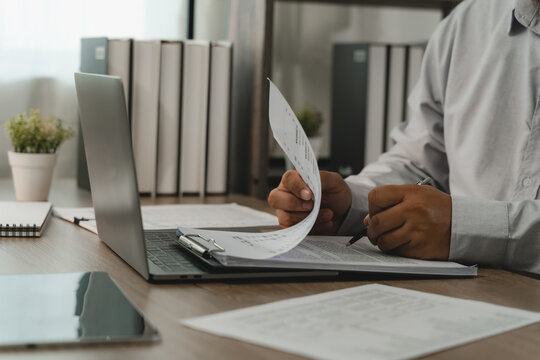 Businessman checking payroll documents on clipboard beside laptop for payroll processing, salary review, employee records, HR administration, accounting workflow.