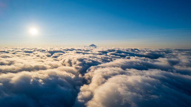 Aerial view of a mountain peak rising above a vast sea of clouds under a clear blue sky with bright sunlight in Bajawa, Nusa Tenggara Timur, Indonesia.