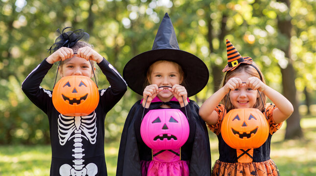 Three children in Halloween costumes holding pumpkin candy buckets outdoors, surrounded by trees and autumn foliage in a park setting