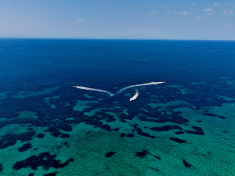 Aerial view of Skala Fourkas Beach with speedboats leaving white wakes on the turquoise and deep blue Aegean Sea in Halkidiki, Greece.