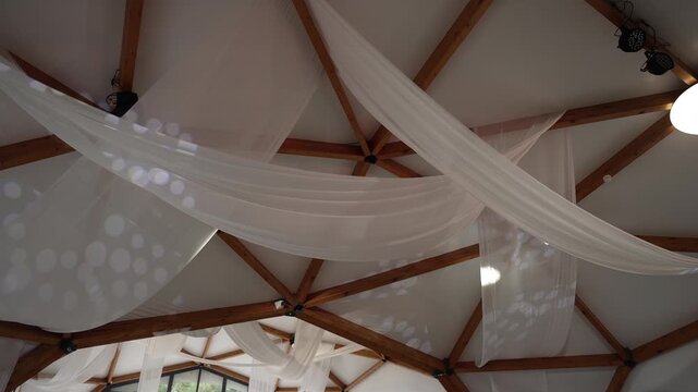 Exposed timber truss ceiling with sheer white fabric swags and shifting decorative light patterns, with fabric, drape, sheer, swag detail, showing ceiling, timber, beam, truss 