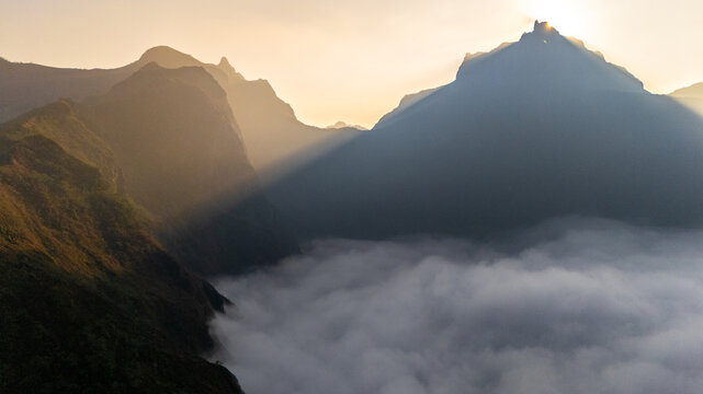 Aerial view of Gunung Kelud at sunrise with dramatic sun rays piercing through the mountain peaks above a thick sea of clouds in Kecamatan Ngancar, Jawa Timur, Indonesia.