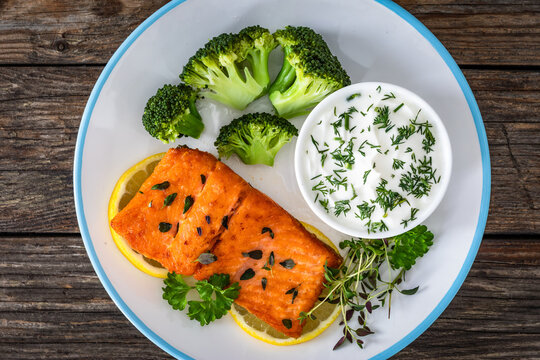 Fried salmon steak with boiled broccoli and dill yogurt sauce on wooden table. Top view
