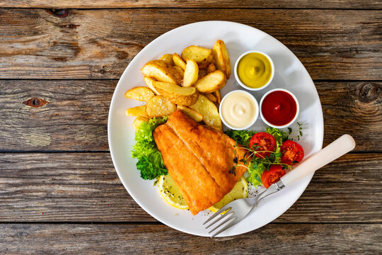 Fried salmon fillet with fried potato wedges, fresh salad and mayonnaise, ketchup, mustard on wooden table. Top view
