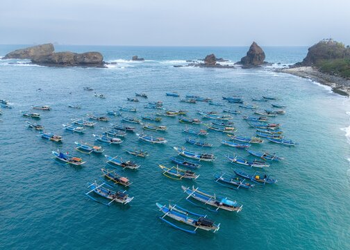 Aerial view of Papuma Beach featuring a large fleet of traditional wooden fishing boats anchored in the turquoise waters near the rocky coastline of Jember, East Java, Indonesia.