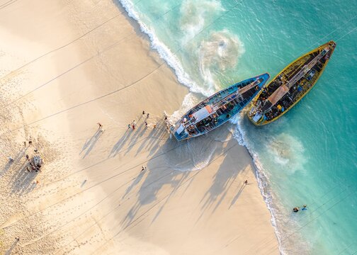 Aerial view of Papuma Beach with traditional fishing boats beached on the white sand, turquoise waves washing ashore, and local fishermen walking nearby in Jember, East Java, Indonesia.