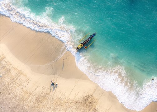 Aerial view of Papuma Beach with a traditional outrigger boat resting on the white sand shoreline, surrounded by turquoise ocean waves in Jember, East Java, Indonesia.