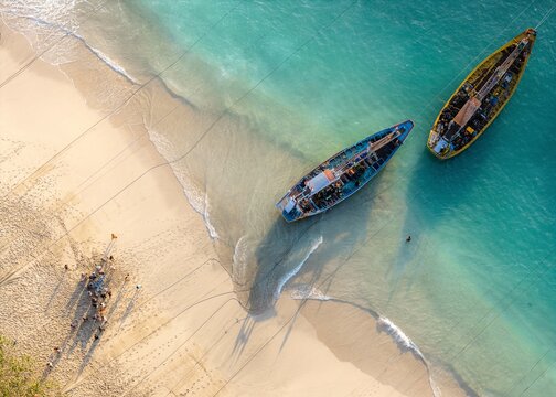 Aerial view of Papuma Beach with traditional fishing boats anchored in turquoise water near the sandy shore and people gathered on the beach in Jember, East Java, Indonesia.