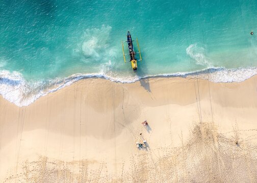 Aerial view of Papuma Beach featuring a traditional outrigger boat resting on the turquoise shoreline with white sand and gentle waves, Jember, East Java, Indonesia.