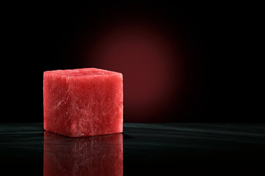 A perfectly shaped red watermelon cube set against a dramatic dark background with reflections. This image works well for creative food and drink advertising concepts.