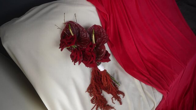 Top-down bouquet on cream cushion with red drapery showing settled tassel repositioning and stem rotation, with bouquet, rose, anthurium, amaranthus detail, showing tassel, drapery, fabric, cushio