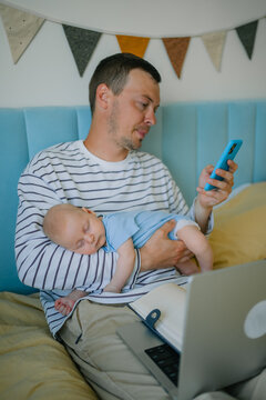 Father multitasking at home, holding sleeping baby while using smartphone and working on laptop, representing modern parenting challenges and work life balance
