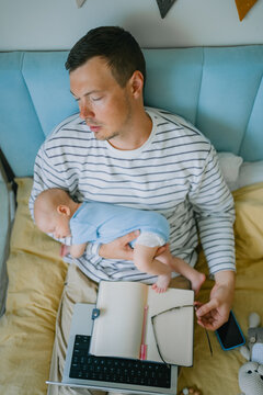Father holding a sleeping baby while appearing tired, simultaneously attempting to work on a laptop and notebook, depicting the challenges of remote work, multitasking, and parental responsibilities