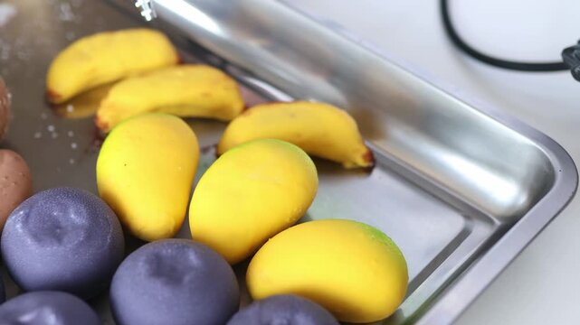 Assortment of colorful fruit-shaped glazed cakes resembling yellow mangoes and purple plums displayed on a metal tray.