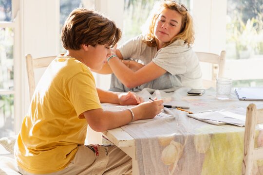 13 year old boy doing homework with older sister looking on in light filled room