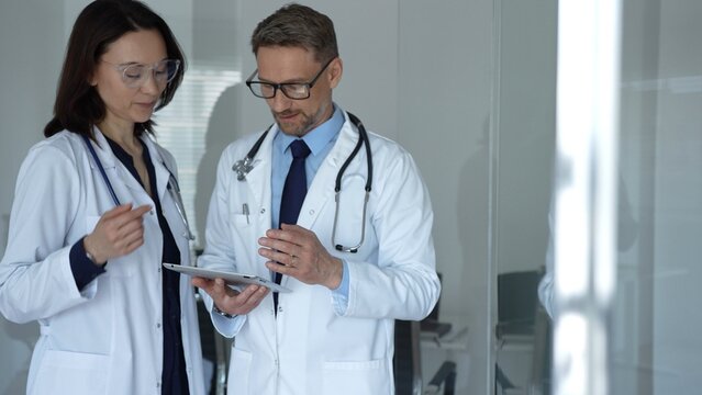 Male and female doctors collaborate over patient data on a tablet in a clinic, both in lab coats with stethoscopes, using digital tech for modern patient care and diagnosis