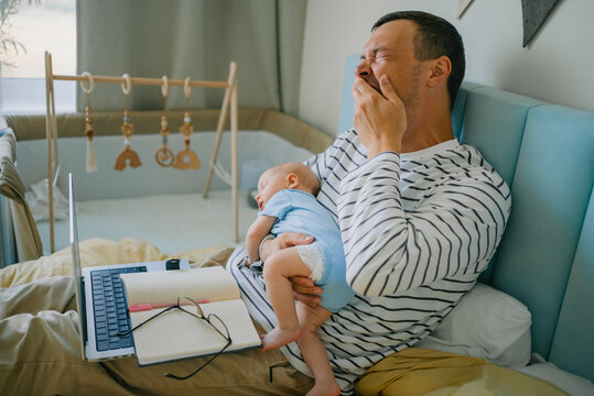 Tired father holding his asleep baby and yawning widely, struggling to stay awake while working remotely from home, navigating the demanding realities of sleep deprivation and new parenting