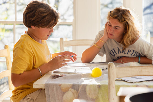 13 year old boy doing homework with older sister looking on in light filled room