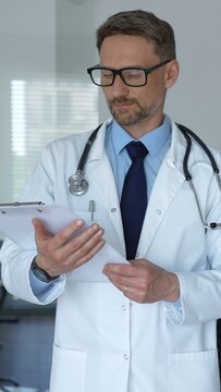 Male doctor in a white lab coat and glasses standing with a clipboard, contemplating healthcare solutions and patient well being in a clinical or hospital setting