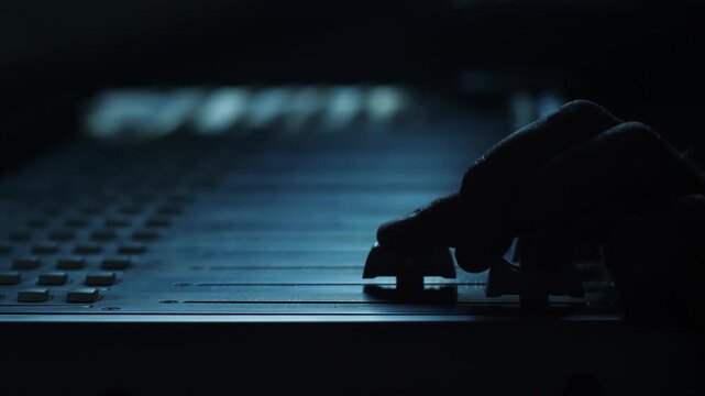 Close-up silhouette of a sound engineer's hand adjusting faders on a mixing console in a dark recording studio. A music producer manually tunes audio channel levels during a professional mixing 