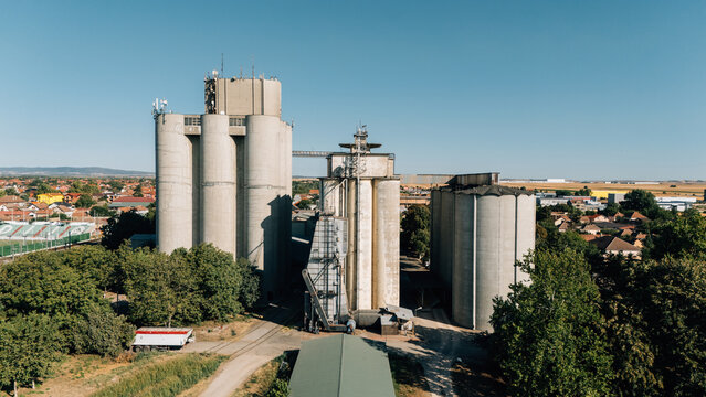Aerial view of industrial grain silos surrounded by green trees and residential buildings in Indjija, Vojvodina, Serbia.