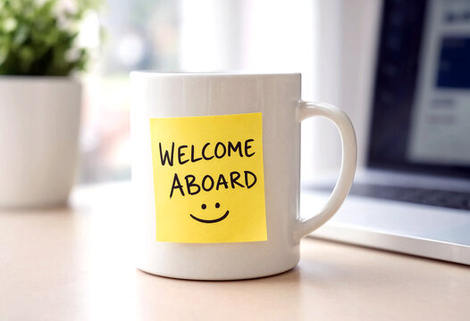 White coffee mug with welcome aboard sticky note on a desk beside a plant and window view
