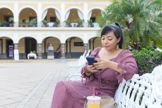 Latina Woman Typing on Smartphone While Sitting on White Bench Outdoors