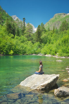 Woman meditates on a rock by transparent turquoise Baduk lake near Dombay under clear blue sky. Alpine Teberda wellness portrait