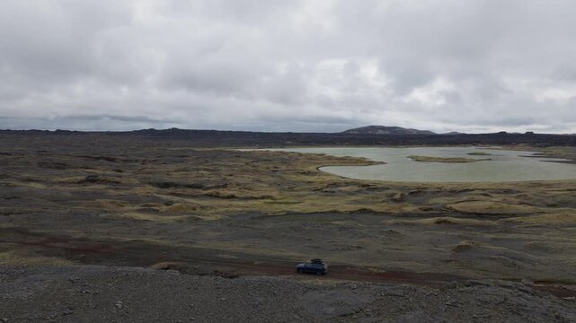 Car travels across barren lava fields beside shallow ponds on Route 425 near Sandvik, Reykjanes, Iceland, within the Mid Atlantic Ridge, captured during a drone tracking shot.