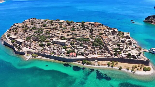 Kalydon ruined settlement grows larger in a close aerial view off Crete, revealing stone foundations, a circular bastion, perimeter wall, trees and dark seabed in turquoise shallows.