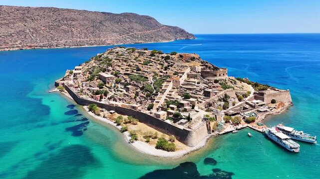 Kalydon fortress rises beside a ferry dock in an aerial view off Crete, with stone ruins, a round bastion, defensive walls, beach shallows and two passenger vessels in bright daylight.