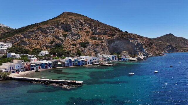 Klima fishing houses line a shallow bay during an aerial view on Milos, with colorful doors, a long stone jetty, anchored boats and rugged cliffs behind the deep blue sea.