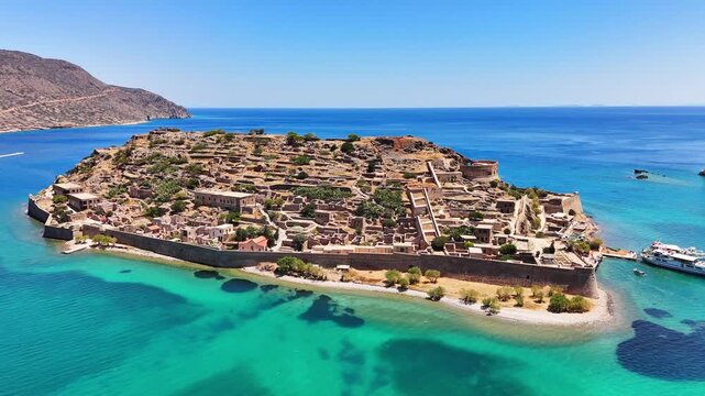 A long wall borders Kalydon fortress during a lateral aerial view off Crete, with stone ruins, a round bastion, narrow beach, dark seabed and a passenger ferry beside turquoise water.