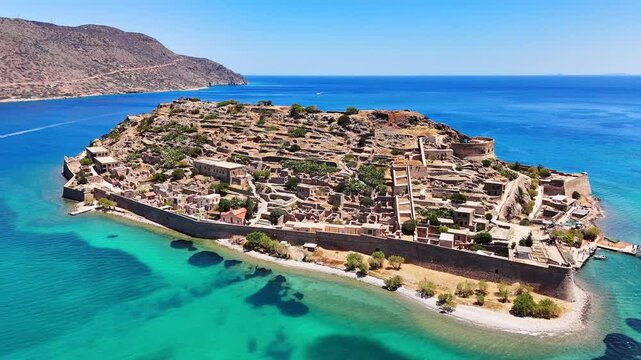 Kalydon fortress walls and dense stone ruins grow larger during a low aerial approach off Crete, bordered by a narrow beach, turquoise shallows and an arid mountain under clear sky.