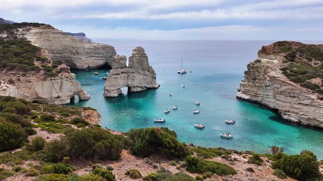 The turquoise channel at Kleftiko opens between sea stacks as an aerial view advances from scrubland on Milos, passing natural arches, anchored boats and a sailboat under soft clouds.