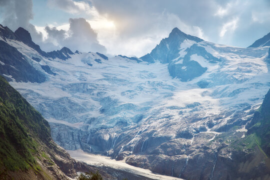 Massive Alibek glacier ice in Dombay flows between rocky cliffs under cloudy sky with sunlight. Caucasian alpine landscape